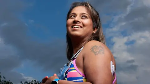 JDRF Asian woman with dark hair and blonde highlights wearing blood sugar sensor on her arm. She is wearing a multicoloured vest top and denim dungarees. The shot is taken side on with her looking past the camera, smiling