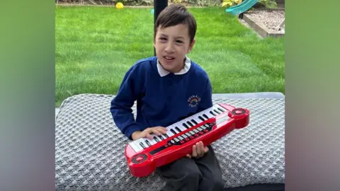 Family A boy wearing a blue jumper and grey trousers. He is holding a children's toy keyboard and there is grass behind him
