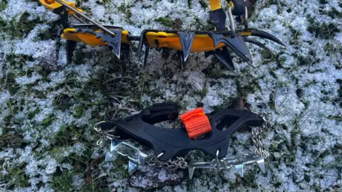 A yellow crampon with sharp black spikes sits on its side, beneath that is a single shoe of microspikes, which have smaller triangular spikes and have a black rubber fixture. There is grass and snow underneath.