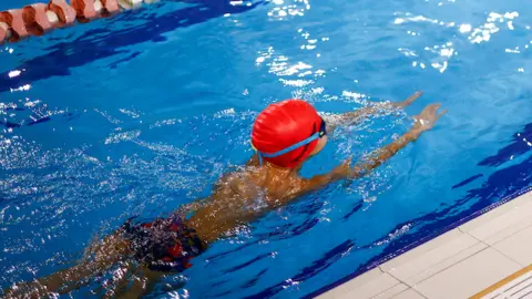 A child wearing goggles and an orange swim hat in the swimming pool in a breast stroke motion.