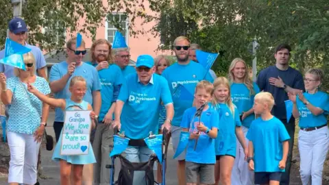 Imogen Treasure-Smith Treasure, walking with his walker and surrounded by family members, from children through to adults. They are wearing Parkinson's UK blue t shirts. 