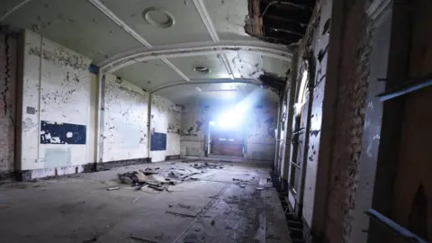 Middlesbrough Council The dilapidated interior of Middlesbrough's Old Town Hall. The room is rectangular-shaped. There are large holes in the ornate ceiling with paint peeling away across each of the walls. The concrete floor is covered with debris. Sunlight is peering through a boarded-up window at the far end of the space.