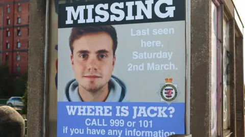BBC A large missing poster for Jack O'Sullivan, close to where he was last seen. The poster features a picture of him staring straight into the camera, in what appears to be a passport or ID picture. He is wearing a light blue hoodie, and has light brown hair. 