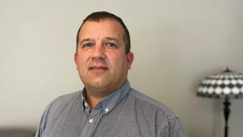 John is at home and this is a head and shoulders shot against a white wall with a black and white lamp beside him. He has no beard in this shot. He was looking into the camera and was wearing a button-down collared shirt.