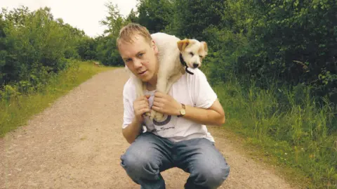 Family photo A man with blonde hair, wearing light blue jeans and a white t-shirt, squats over a path. He has a small white dog with light brown ears around his neck, and he is holding the dog's legs. The dog wears a black collar with a silver tag. The man and the dog are on a path, which is surrounded on both sides by grass and large green bushes.