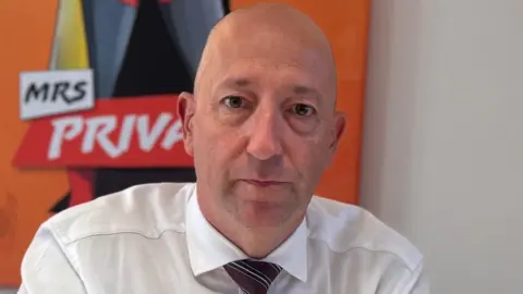 Paul Vane sitting at a desk with his hands clasped. He has a serious expression, and is wearing a white shirt and stripey tie. He is bald. There are campaign posters visible in the background.