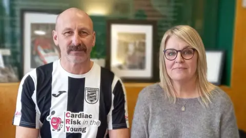 Stephen Huntley/BBC A head and shoulders image of Trevor Chenery and Jane Wilson. Trevor is wearing a black and white striped Halesworth football kit while Jane has a grey jumper and glasses on. They are sitting next to each other and both looking into the camera. In the background there are blurred photo frames. 