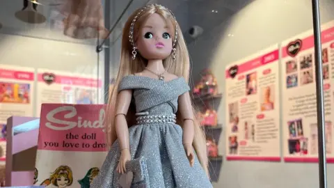 A doll with blonde hear wearing a silver glittery dress and headband