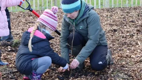 A young girl in blue wellies with a pink butterfly pattern wearing a pink and white striped woolly hat, navy coat and blue leggings plants a tree in a park with a women wearing a blue and green striped woolly hat green coat, blue trousers and wellies helping her dip the tree. 