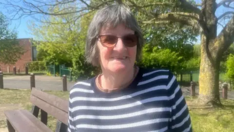 Andrew Turner/BBC Gill Baker is a woman with short grey hair, who is looking into the camera and smiling. She is wearing sunglasses and a navy and white striped jumper. It has been taken on a sunny day, and behind her there is a bench, a tree, and a path, which is lined by wooden bollards.