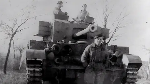 Richard Aldred A black and white photo showing Richard Aldred standing in army uniform in front of a tank. On the tank are two other men also wearing army uniform. There are a few slim trees behind them.