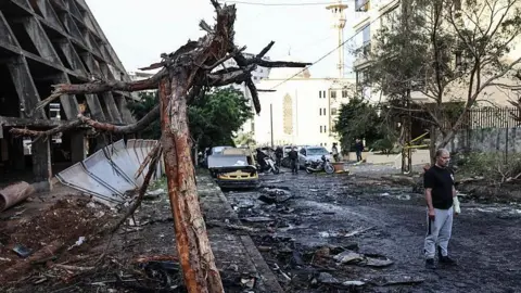 A man stands in the street next to a building and tree which are both heavily damaged and blackened. Rubble lines the street and cars in the background are also damaged.