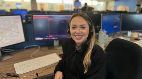 Rhiannon Gardener, wearing a black fleece and headset, sits at a desk in the police control room