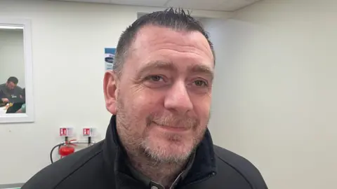 A man, pictured from the shoulders up, with short black hair and stubble. He is wearing a black jacket. 