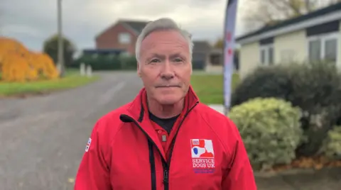 A man with short grey hair faces the camera - he's wearing a bright red jacket with the words Service Dogs UK written on it in white.