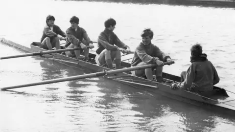 Nick Cooper A black and white photo of a group of five young boys rowing on a body of water.