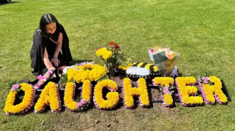 Scarlett Black Mrs Black at a graveside with a large yellow floral wreath spelling out the word 'daughter'