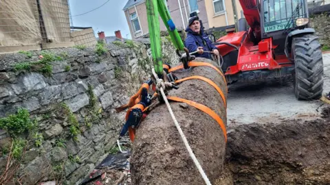 PO Paul Cartwright overseeing the safe lifting of the bomb. Straps have been tightened around the bomb to lift it out of the ground. Mr Cartwright is standing behind it. A stone wall and houses surround the area.