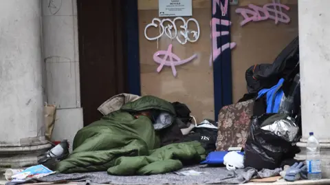 PA Media A rough sleeper in a doorway, wrapped in a green sleeping bag, surrounded by their belongings and a pigeon