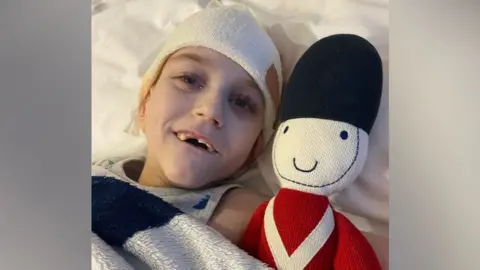 Toby laying in bed next to a knitted toy soldier. He has a bandage on his head and is wearing a grey vest. He is smiling at the camera.
