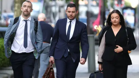 PA Media Daithí McKay is walking along a street with another man and a woman. Mr McKay has short black hair and a goatee beard. He is wearing a light blue jacket and a blue tie with a white shirt and dark trousers. The man and women to his left are professionally dressed and carrying brief cases and pulling a baggage trolley.