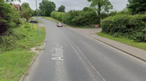 Google Allum Lane when it is not flooded. It shows a signpost pointing right towards the entrance of a recycling centre. A red car is driving down the hill.