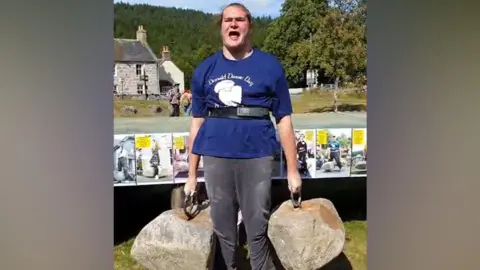 Leigh Day Solicitors A tall, heavily built man in his late teens, with long brown hair tied back, is standing straight lifting two large boulders which have had metal rings bolted into them. He is wearing a blue t-shirt with a slogan reading "Donald Dinnie Day" and his mouth is open in a roar. 