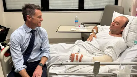 Reuters Mr al Ahmed is lying on a hospital bed facing left. His left arm is bandaged and he is wearing white. At the end of the bed, sits Mr Minns who is talking to him. They are in a hospital room