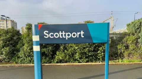 A blue sign for Stockport station, re-written to say Scottport in white lettering, erected outdoors on a train station platform. 