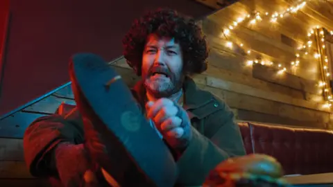 A man with a beard and large curly brown hair sits on a red, leather bench and looks at a spike through his shoe. 