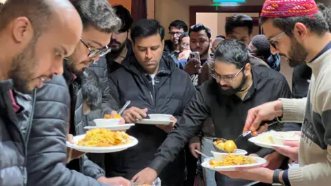 Lots of men are around a table and they're reaching down to ladle food onto their plates.