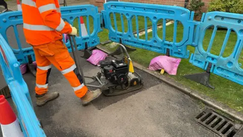 Chris McHugh/BBC A workmen holds a large piece of machinery over the top of a filled pothole. Blue barriers can be seen around the outside and a grass front lawn of a nearby house can be seen