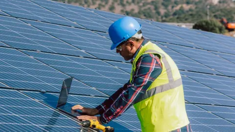 A man wearing a high vis jacket and hard hat operates a computer resting or an array of rooftop solar panels