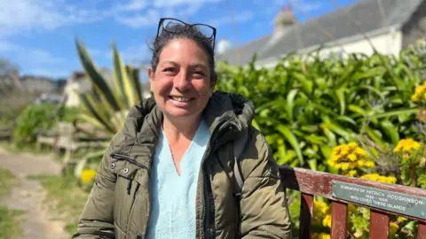 Jenny sat on a park bench, she is wearing a light blue jumper and a green coat while smiling at the camera.
