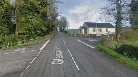 A crossroads at the Glenavy road and whinney hill. On the corner of the road on the right of the picture is a white church hall. The T-junction on the left has green trees on the corner 