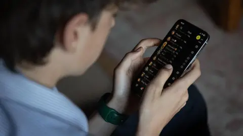 A photo illustration showing a teenage boy looking at his phone as he scrolls through the Snapchat app. He is wearing a watch on his left wrist, and has short black hair. 