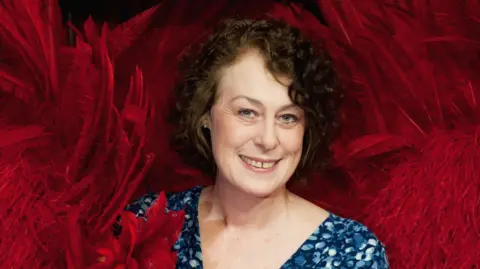 A glamorous-looking lady with short, curly brown hair and wearing a blue dress. She is surrounded by red feathers all around her. She is smiling into the camera. 