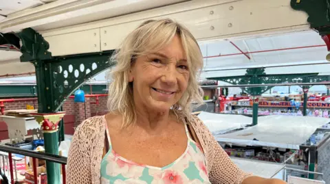 BBC Linzy has shoulder-length bloned hair with a fringe. She is pictured smiling and looking into the camera. She's wearing a blue top with pink flowers and a white knitted cardigan. She's stood on am upstairs mezzanine of an indoor market, which has green wrought iron pillars and a white roof. Beyond Linzy you can see the tops of the market stalls.