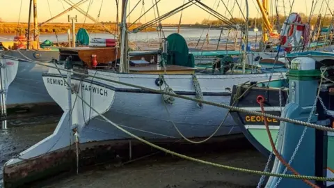 There are a number of boats moored. The background weather is clear and looks like it could be a sunrise or a sunset.
