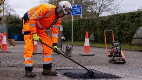 A man, in yellow high-vis, fixing a pot hole in a road, leaning over, holding a broom. Equipment is around him and traffic cones. He has on gloves, boots, a helmet and googles. A bush it to the right and a sign. 
