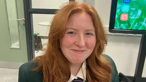 A girl with long ginger hair is smiling and looking into the camera. She is wearing a green school blazer, a tie and a white shirt.