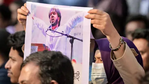 Getty Images A woman wearing a face mask holds up a photo of Sharif Osman Hadi, senior leader of the student protest group Inqilab Mancha