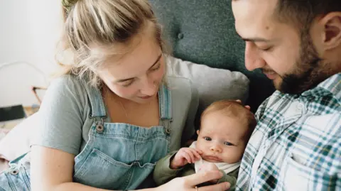 Mum and Dad both look down at their newborn baby and Mum holds his tiny fingers in hers