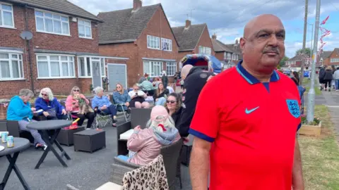 Kash is wearing a red England football top and is stood in a street with people sat in chairs in the street behind him.