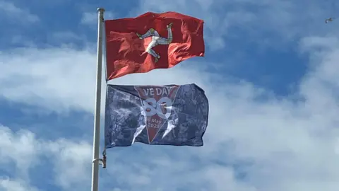 A red Manx flag with a yellow and white triskllion flies on a flag pole. Underneath there is a blue VE Day 80 flag. The sky behind is blue with white clouds.