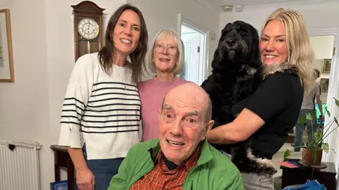Ceri Evans A family photo. A woman, in a white and black stripped top, wearing jeans, next to a woman with bobbed grey hair, and glasses, by a woman, with long blonde hair, holding a black dog, wearing a black top. A man is sat down in front, they are all smiling. They man is wearing a checked shirt and green jumper. They are in a living room with a grandfather clock to the left and a plant to the right. 