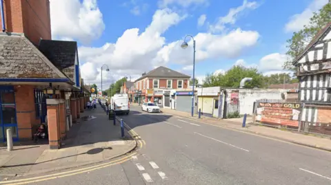 A generic photo of Bilston High Street, captured at a junction. The high street goes from the bottom right towards around the middle of the image. Buildings are on both sides of the road and a white car and van are in the background.