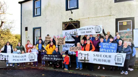 Local residents of Brydekirk holding banners reading "Save our pub" outside of the Brig Inn. The people are of a range of ages and smiling towards the camera. 