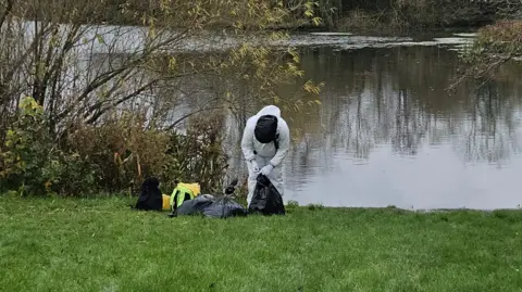 Curious Critters Person wearing white suit stood near lake carrying a black plastic bag. He is stood near to a tree and you cannot see their face.