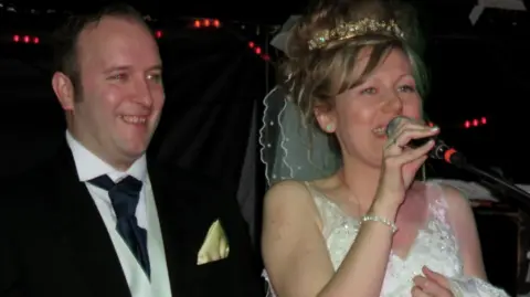 A man in a suit and a woman in a wedding dress are standing on a stage with lights behind them. The bride is holding a microphone.
